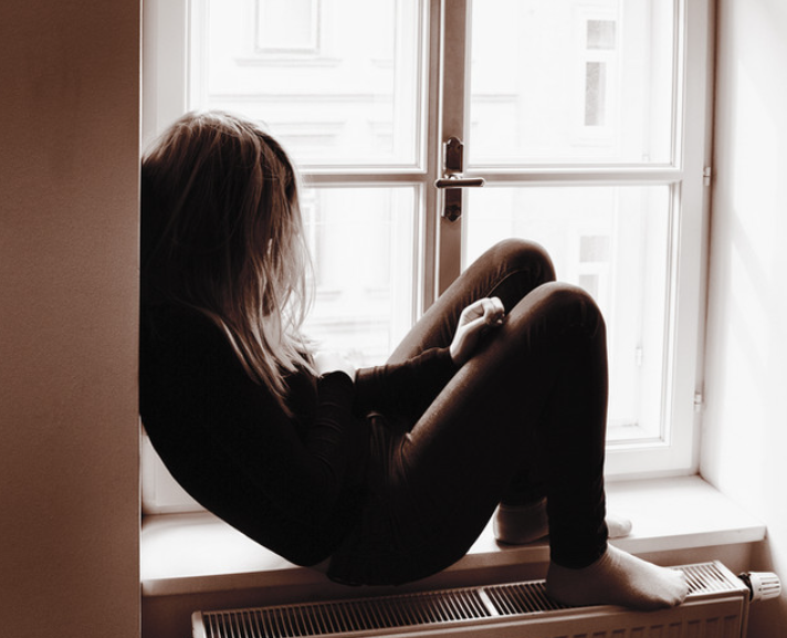 Woman sitting by a window, curled up with her knees drawn to her chest, appearing deep in thought or emotionally distressed